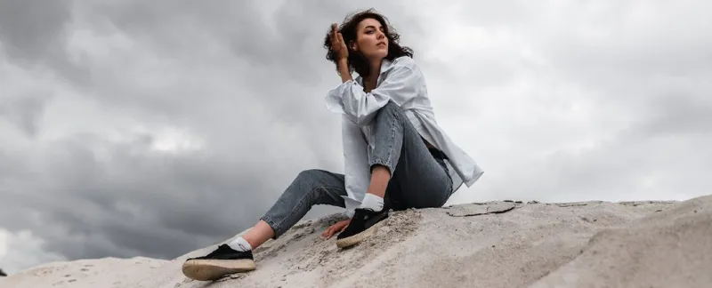 Woman sat on sand ridge wearing a shirt and jeans with dark cloudy skies above